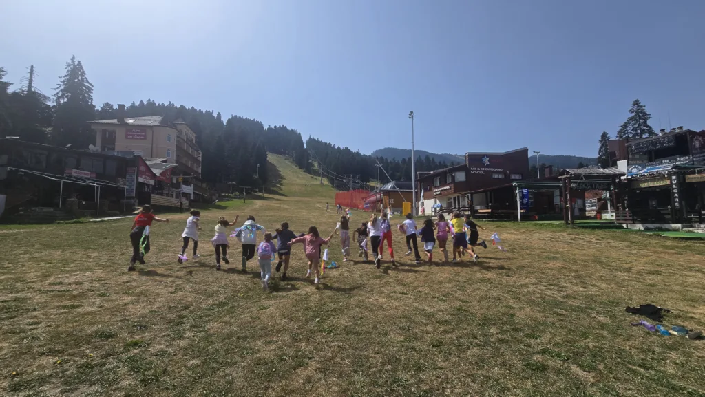 Група деца тичат по широка поляна в планината